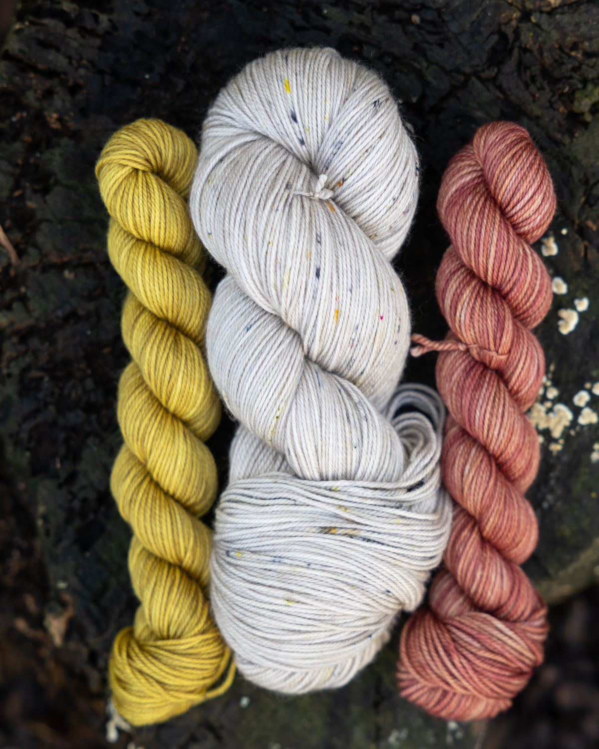 Full skein of light ecru colored yarn with a mustard colored quarter-skein on one side and a terracotta orange colored quarter-skein on the other side