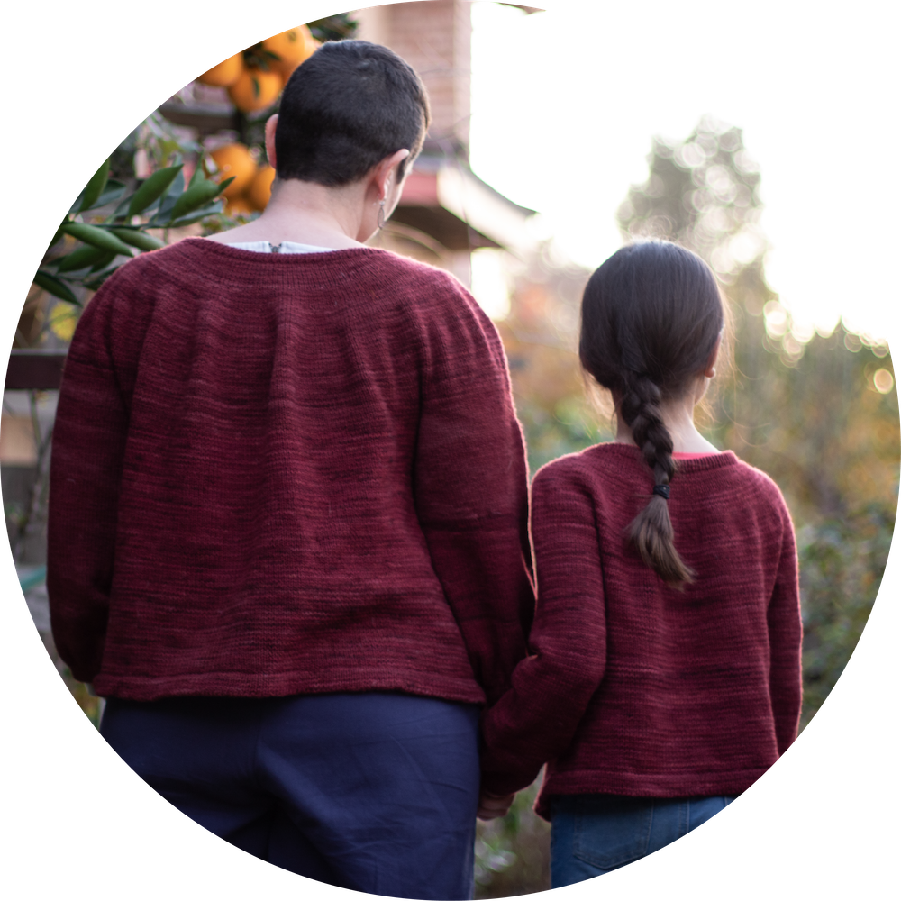 Parent and child are standing, backs to the camera. They are wearing matching deep red-colored handknit cardigans.